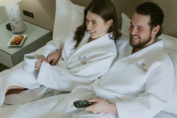A couple lay on the bed in their comfortable embroidered Bayside waffle robes
